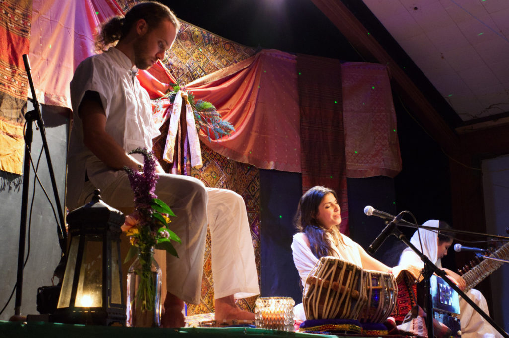 Yogic singer on stage with female sitar player to her right, male tabala player to her left on a chair
